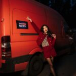 Asian woman in red attire posing confidently by an orange news van at night.