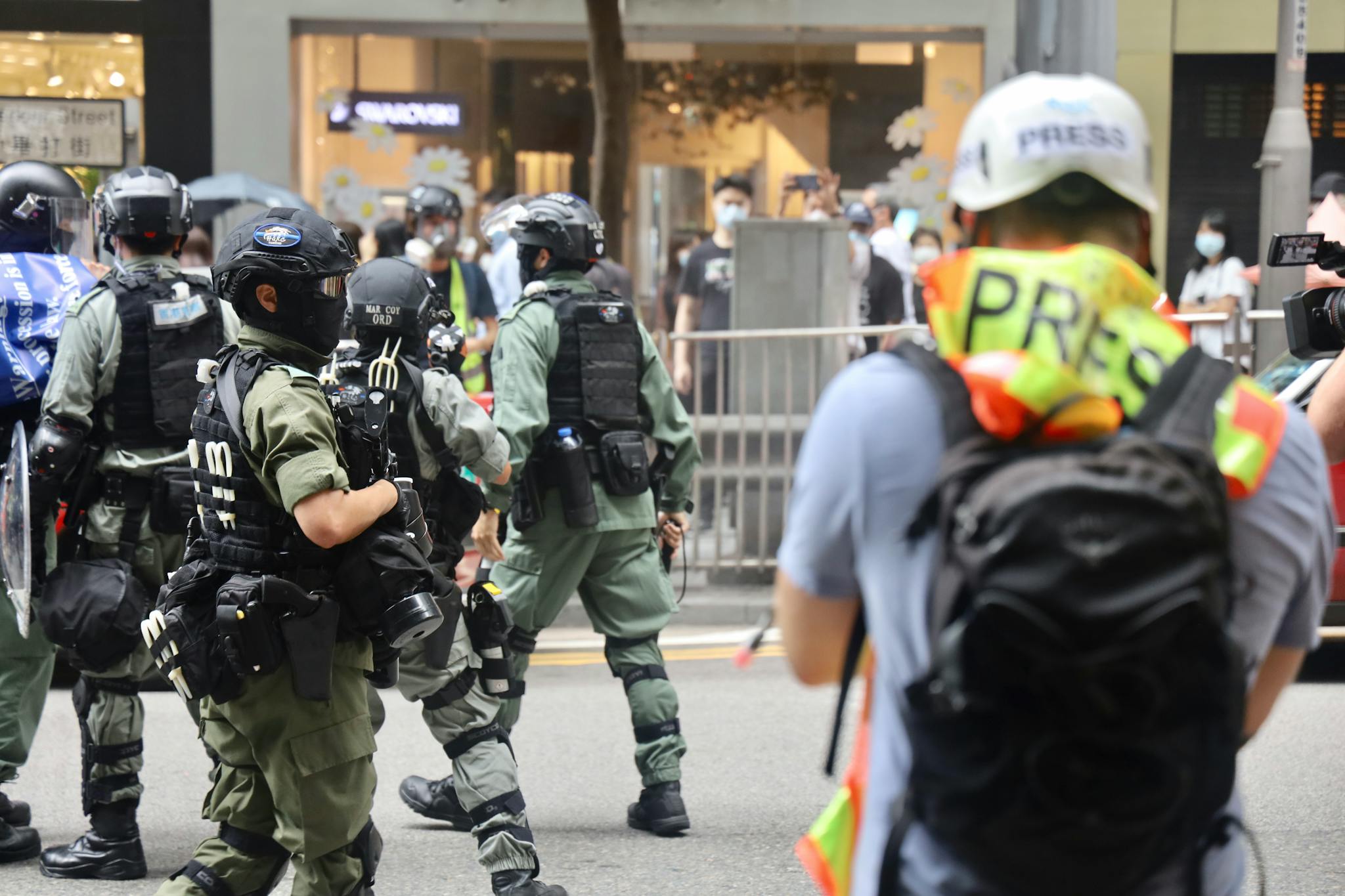 Riot police and journalists during a city protest, wearing protective gear.